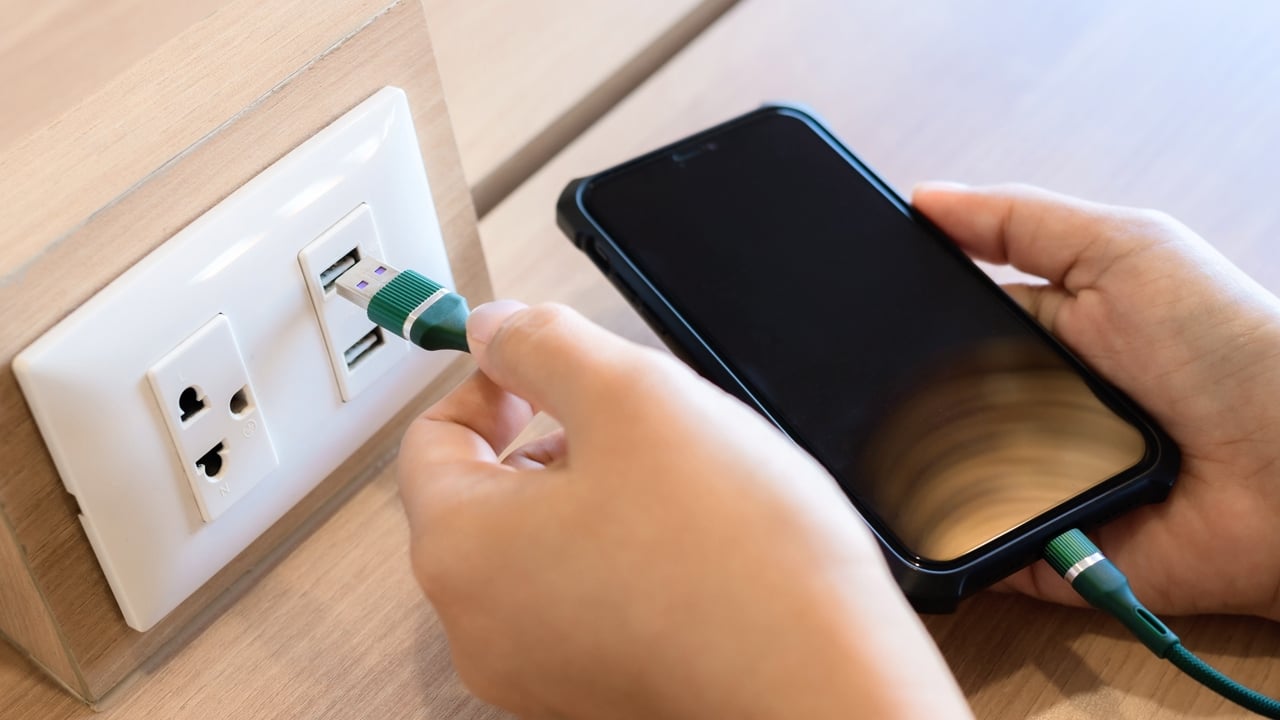Person's hand is plugging a phone charger into a USB socket built into the desk.