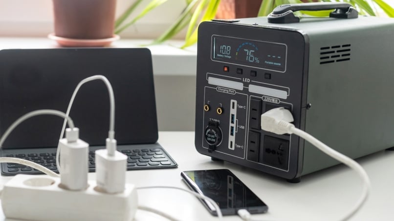 Portable power station with digital battery display powering a laptop and charging mobile devices via USB and AC ports on a desk.